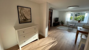 Ein Zimmer mit Holzboden, einem weißen Schminktisch, einem Gemälde, einem Fernseher, einem Esstisch und einem Fenster mit Blick auf Grünflächen.
