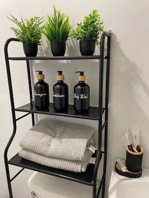 Ein schwarzes Metallregal mit drei Blumentöpfen, drei beschrifteten Flaschen mit Shampoo, Conditioner und Duschgel sowie einem gefalteten Handtuch.