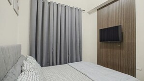 Une chambre à coucher avec un lit, des rideaux gris, un téléviseur fixé au mur et un mur recouvert de panneaux de bois.