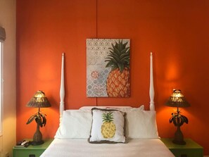 Een slaapkamer met een bed, twee nachtkastjes, een aan de muur hangend schilderij van een ananas en twee aan de muur bevestigde lampen.