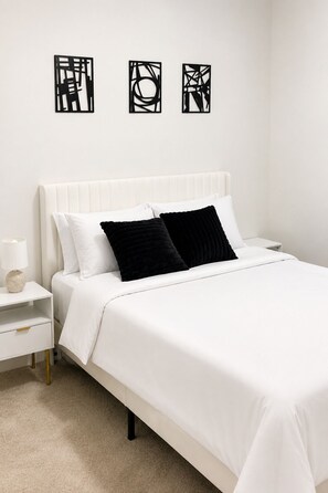 Ein Schlafzimmer mit einem weißen Bett, zwei schwarzen Kissen, einem weißen Nachttisch, einer Lampe und drei abstrakten Wandbildern.