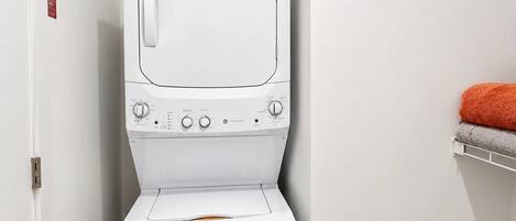 A laundry room with a stacked washer and dryer, a door with a red sign, and a shelf with a towel.