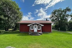 Ein rotes Holzgebäude mit weißer Tür und Fenstern, umgeben von grünem Gras und Bäumen.
