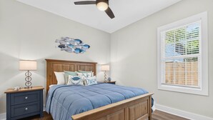 A bedroom with a wooden bed, two bedside tables with lamps, a ceiling fan, a wall art piece, and a window with blinds. - Spacious 4 BDR/3 BA pet friendly <10 min walk to beach + Hot Tub (Oak Island)