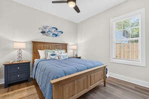 Ein Schlafzimmer mit einem Holzbett, zwei Nachttischen mit Lampen, einem Deckenventilator, einem Wandbild und einem Fenster mit Jalousien.
