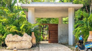 Uma trilha de pedras leva a uma construção de concreto com porta de madeira, cercada por plantas tropicais e uma grande rocha com a inscrição 'SEREMONIA HABITAT'.