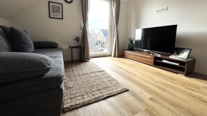Ein Wohnzimmer mit Sofa, Fernseher, Holzboden und einem Fenster mit Vorhängen.