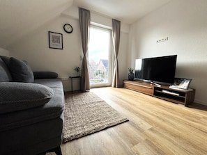 Ein Wohnzimmer mit Sofa, Fernseher, Holzboden und einem Fenster mit Vorhängen.