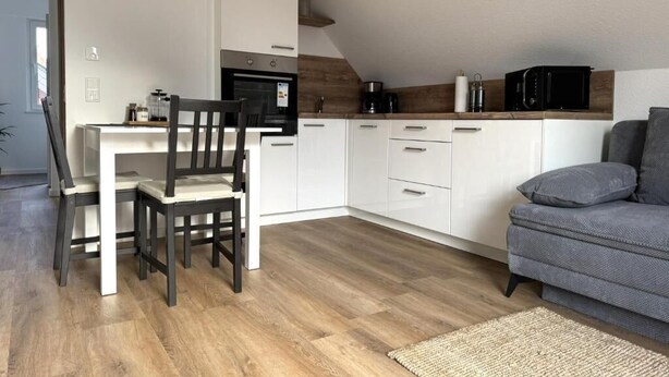 Eine kompakte Küche mit weißen Schränken, einem Esstisch mit Stühlen, einem grauen Sofa und einem Holzboden.