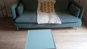 Ein blaugrünes Sofa mit einem gemusterten Kissen und einer weißen Decke, ein kleiner blauer Tisch und ein Holzboden.