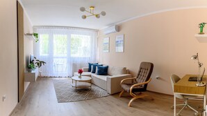 Una sala de estar moderna con un sofá, una butaca, una mesita y un escritorio con su silla. Cuenta con un ventanal con cortinas traslúcidas, una lámpara de techo y aire acondicionado instalado en la pared.