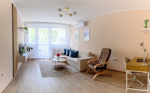 Un salon moderne comprenant un canapé, un fauteuil, une petite table et un bureau avec une chaise. Il y a une grande fenêtre avec des rideaux légers, un luminaire au plafond et un climatiseur fixé au mur.