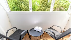 Un balcón con dos sillas y una pequeña mesa, un libro sobre la mesa, y vistas a árboles a través de la ventana.