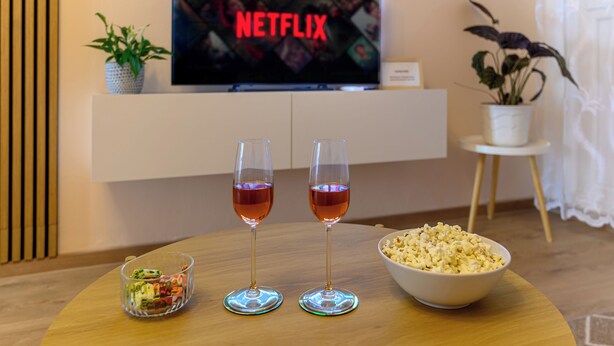 Una sala de estar con un televisor mostrando el logotipo de Netflix, dos vasos de líquido rojo, un cuenco de palomitas de maíz y un pequeño recipiente de vidrio sobre una mesa de madera.