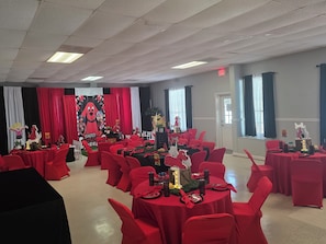 Una habitación con mesas dispuestas para una celebración, con manteles rojos y sillas, un cartel grande con un personaje de dibujos animados, y una puerta con interruptor de luz.