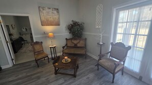 Een woonkamer met een houten vloer, een ronde salontafel, twee fauteuils, een bijzettafeltje met een lamp, een plant in een pot en een raam met dunne gordijnen.