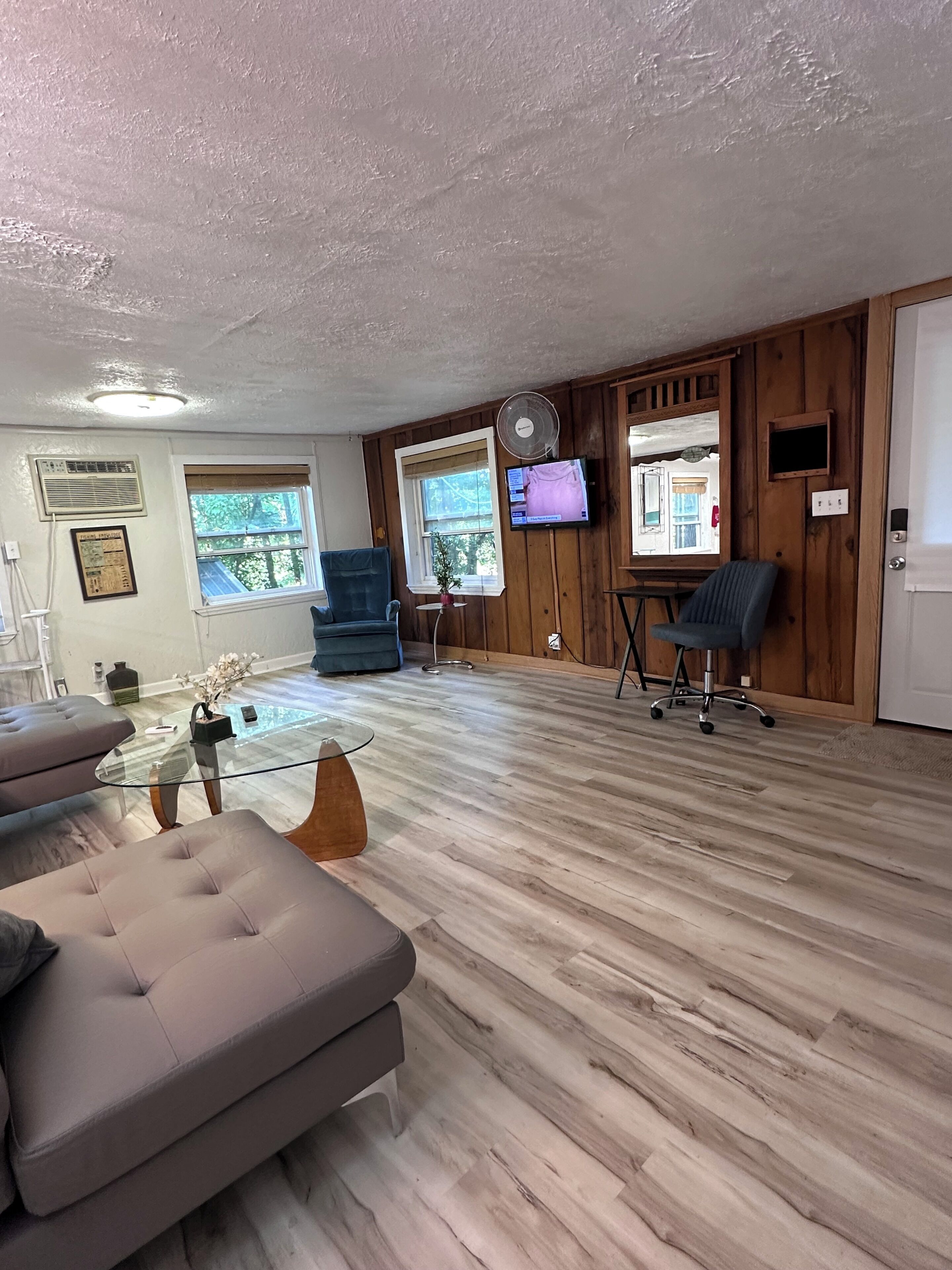 Une pièce spacieuse avec un plancher en bois, une table basse en verre, un canapé, un téléviseur fixé au mur et une fenêtre donnant sur des arbres.