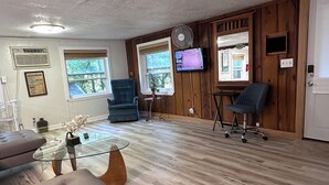 Ein geräumiger Raum mit Holzboden, einem Glastisch, einer Couch, einem an der Wand montierten Fernseher und einem Fenster mit Blick auf Bäume.