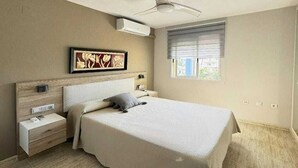 Une chambre à coucher comprenant un lit, des tables de chevet, un ventilateur de plafond, un climatiseur, une fenêtre avec des stores et une œuvre d’art accrochée au mur.