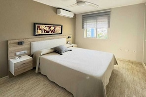 Une chambre à coucher comprenant un lit, des tables de chevet, un ventilateur de plafond, un climatiseur, une fenêtre avec des stores et une œuvre d’art accrochée au mur.