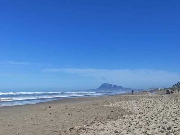 Ein Strand mit einem Berg im Hintergrund, sandigem Untergrund und blauem Himmel.
