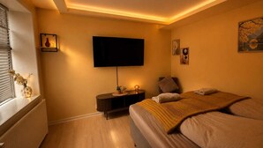 A bedroom with a bed, a bedside table, a TV mounted on the wall, and a window with blinds.