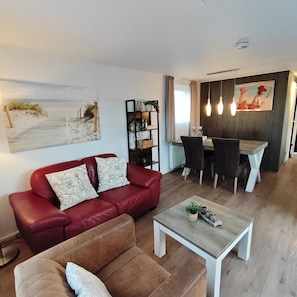 A living room with a red sofa, a brown armchair, and a coffee table. There is a dining area with a table and chairs, and a painting on the wall. - Modern 4-person chalet in Zeeland—near the beach, Veere, Middelburg, and Domburg (Serooskerke)