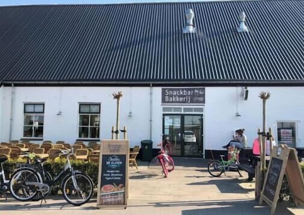 A building with a sign reading 'Snackbar Bakkerij.' There are bicycles parked in front, and a person is visible inside. - Modern 4-person chalet in Zeeland – quiet location near Domburg and Middelburg (Serooskerke)
