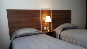 Habitación de hotel con dos camas, cabecero de madera, mesita de noche con lámpara y otro mueble auxiliar.