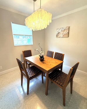 A dining room with a wooden table, six chairs, a hanging light fixture, a window with blinds, and a wall-mounted artwork.