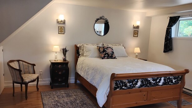A bedroom with a wooden bed, a nightstand, a chair, a mirror, and a window with curtains. - Victoria Garden Suite (Victoria)