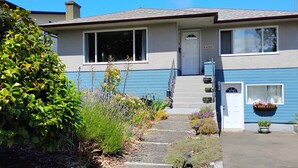A two-story house with a front porch, a visible door, and a small garden area with plants and a pathway leading to the entrance. - Victoria Garden Suite (Victoria)