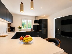 A modern kitchen with black cabinets, a central island with a fruit bowl, a built-in oven, and a water dispenser. - FAMILY VILLA 190 m² (190 sq.m.) - UP TO 14 PEOPLE - GARDEN 2 500 m² - BEACHES 5 min away (Vendays-Montalivet)