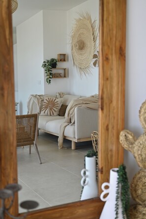 Ein Zimmer mit einer Couch, einem Korbstuhl, einem Spiegel und Dekorationselementen, darunter ein Wandbehang aus Bändern und eine Pflanze.