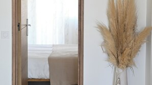 Uma porta que dá para um quarto com uma cama, um lustre pendurado e um vaso alto com plumas de capim-do-avelar.