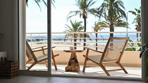 Uma varanda com duas cadeiras de vime e uma mesa de madeira, com vista para uma praia com palmeiras e o oceano.