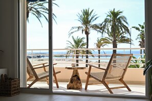 Ein Balkon mit zwei Korbstühlen und einem Holztisch mit Blick auf einen Strand, Palmen und das Meer.