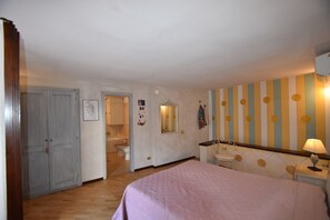 Ein Schlafzimmer mit einem Bett, einem Nachttisch mit Lampe, einem Spiegel und einem im Hintergrund sichtbaren Badezimmer.