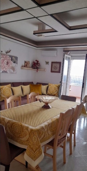 Un comedor con mesa y sillas, un sofá con cojines, un elemento decorativo en la pared, y una puerta que da a un balcón exterior.