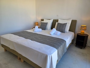 Ein ordentlich bezogenes Bett mit weißer und grauer Bettwäsche, zwei Nachttischlampen und ein Nachttisch aus Holz.