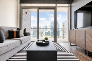 A modern living room with a sofa, coffee table, and a view of the cityscape through large windows. - Skyline Views from the High Floor | Walk to AAC (Dallas)