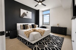 A modern bedroom with a large bed, a ceiling fan, a window with a city view, and a dark dresser.