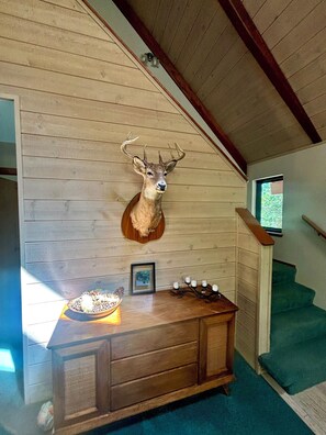Una habitación con pared de madera, cabeza de ciervo montada, aparador de madera, un cuadro enmarcado y una ventana.