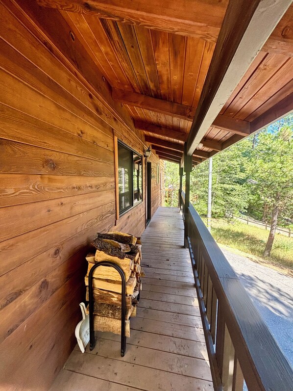 A wooden porch with a railing, a stack of firewood, and a view of trees.