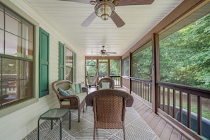 Eine überdachte Veranda mit Korbmöbeln, einem Deckenventilator und großen Fenstern mit Blick auf viel Grün.