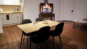 Een eetruimte met een houten tafel, zwarte stoelen, een televisie waarop een haard wordt getoond, en een keuken met witte kasten.