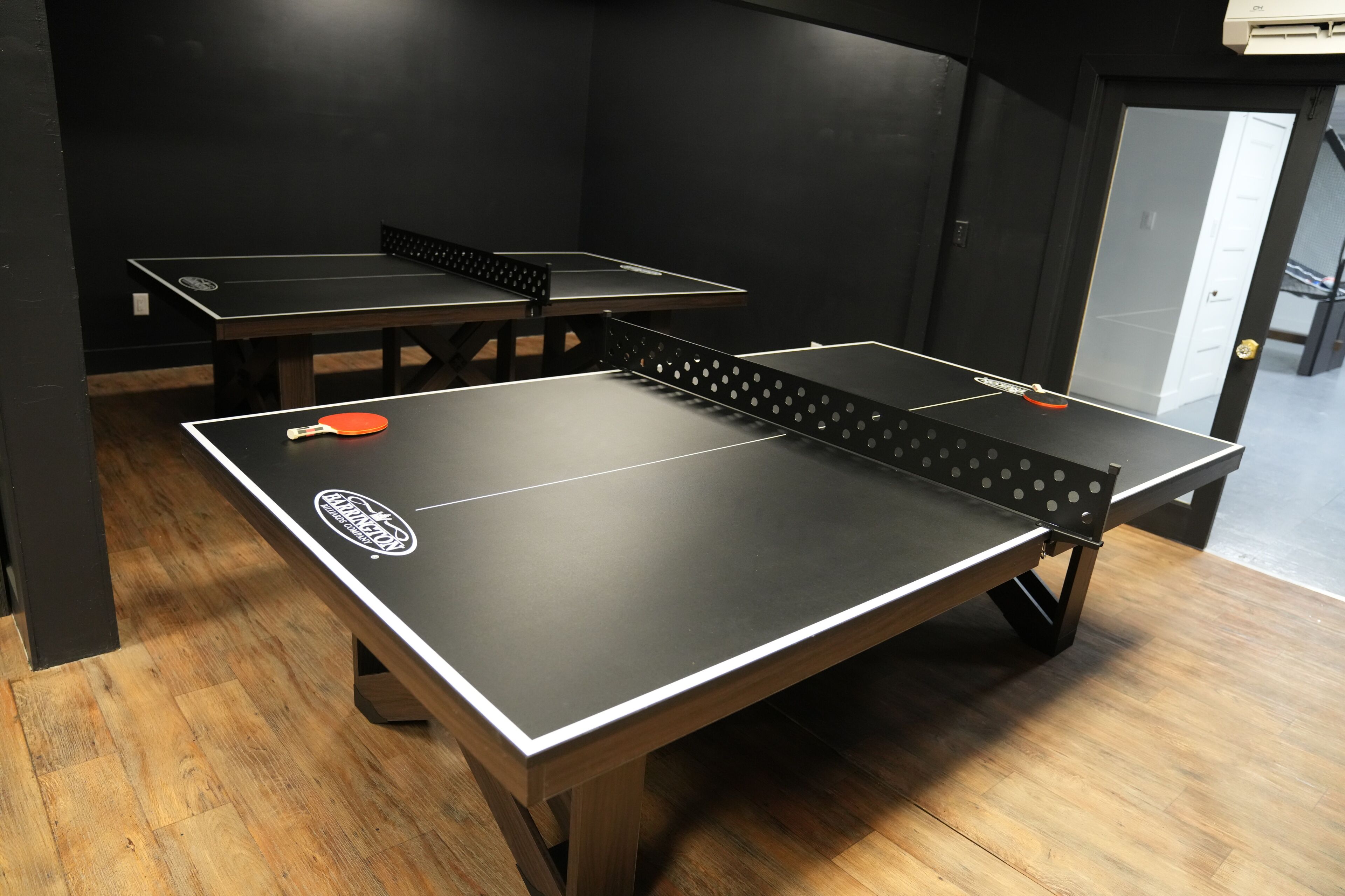 Une pièce équipée de tables de ping-pong, dotée d’un plancher en bois et d’une porte donnant accès à un autre espace.