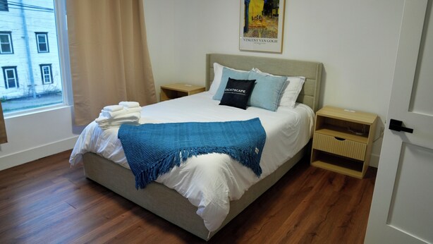 Een net opgemaakt bed met een blauwe plaid, een kussen met tekst en een kunstwerk in een lijst aan de muur.