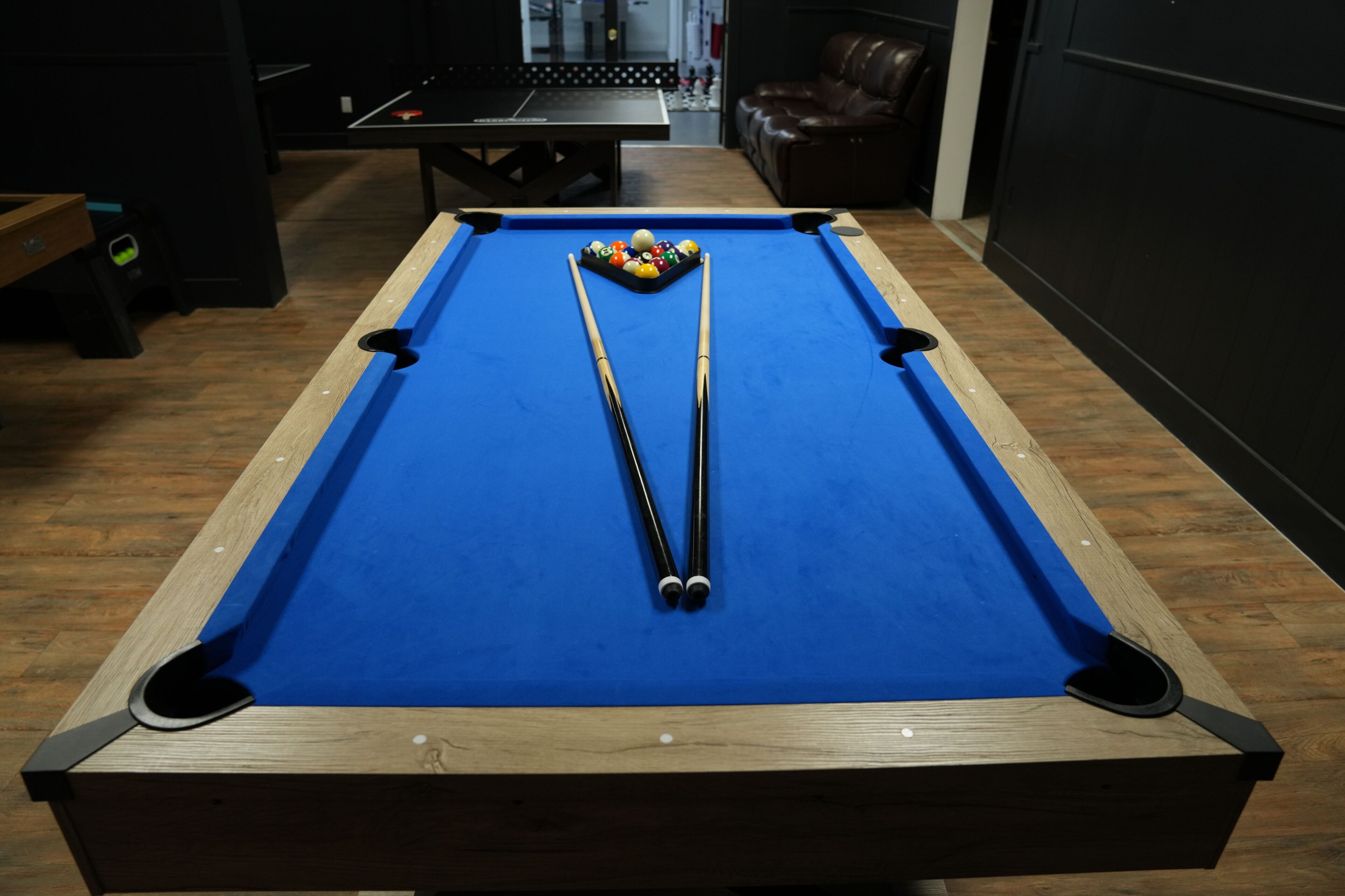 A pool table with balls and cues, a wooden floor, a leather couch, and a ping pong table in the background.