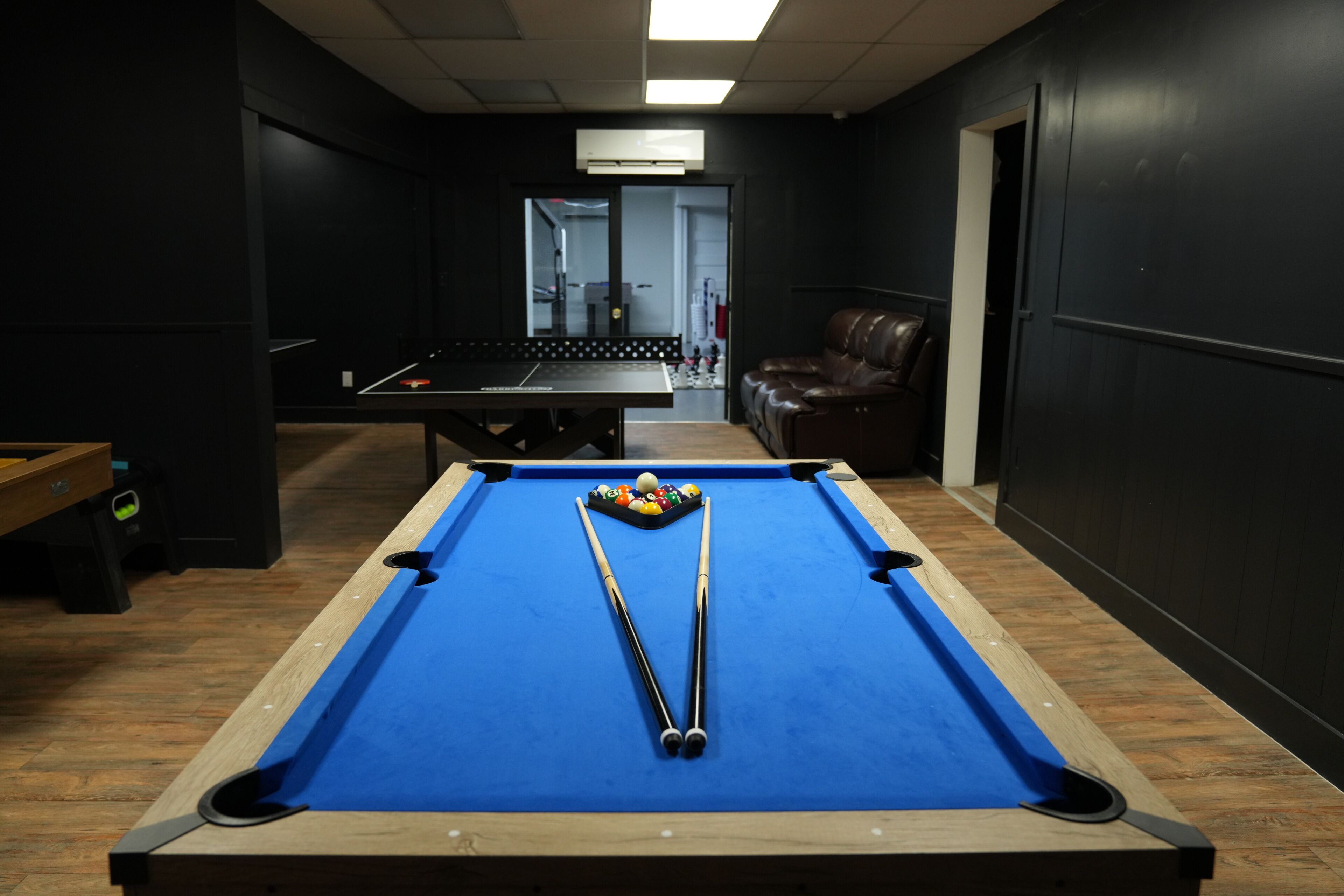 Une table de billard avec deux queues et des billes, un fauteuil en cuir et un banc en bois, dans une pièce aux murs sombres et dont la porte est visible.
